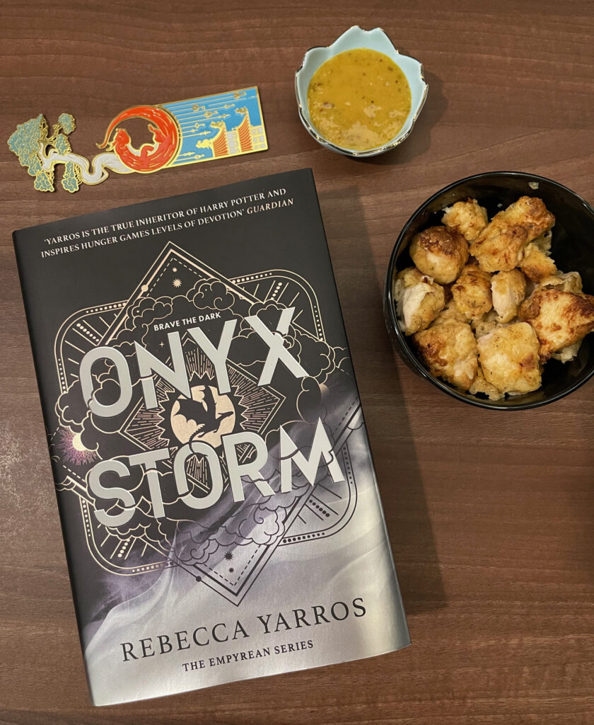 Physical hardcover version of ONYX STORM, photographed with a bookmark and a bowl of chicken nuggets as a snack